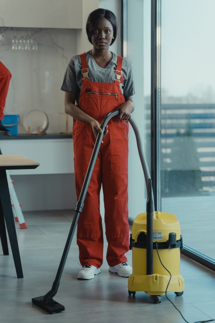 gallery-3 An adult woman in red coveralls operates a vacuum cleaner in a modern indoor setting, focusing on cleaning tasks.