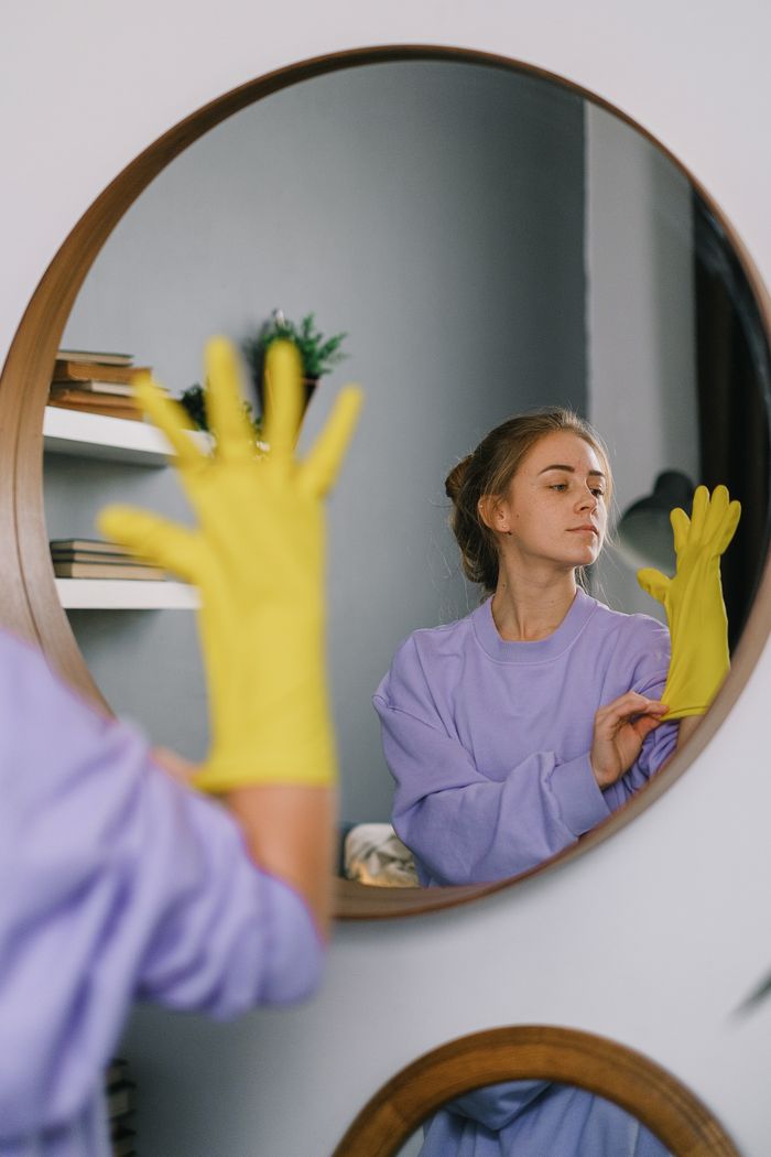 about-01 A woman wearing yellow gloves is reflected in a wall mirror indoors, exuding a serene expression.